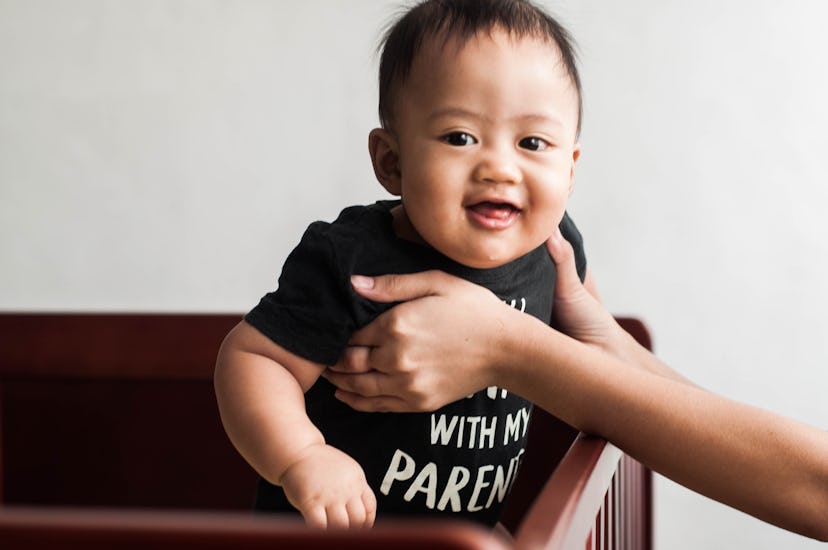 A waist-up portrait of a Southeast Asian baby boy walking on the crib with assistance of his mother