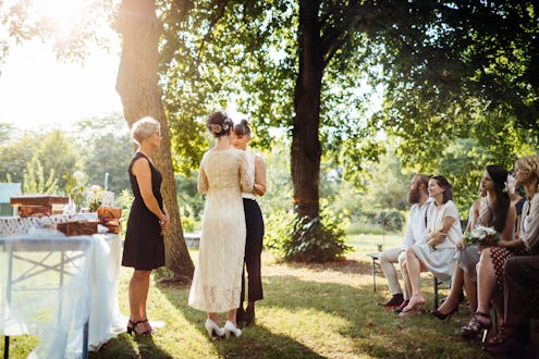 Lesbian couple holding hands in front of the altar. The wedding is outdoors and guest are seated in ...