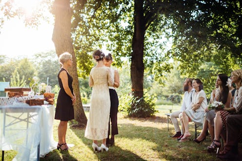 Lesbian couple holding hands in front of the altar. The wedding is outdoors and guest are seated in ...