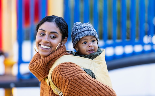 A mid adult woman in her 30s with her 7 month old baby boy on a playground. He is on her back in a baby carrier and mother is looking over her shoulder at him. They are mixed race Hispanic, African-American and Native American.
