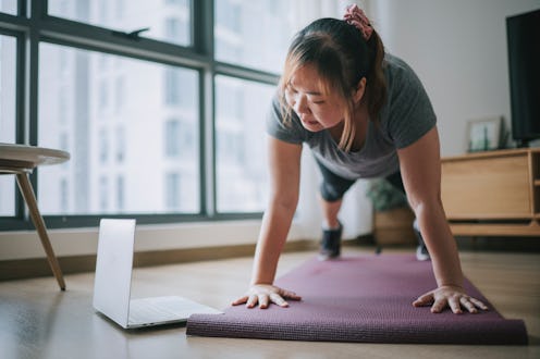 A person looks at their computer while in pushup position on a yoga mat at home. Strength training w...