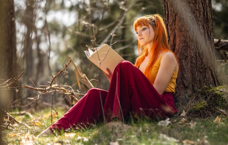 Free spirited young woman outdoors reading a book against a tree in nature, sitting in the park or g...