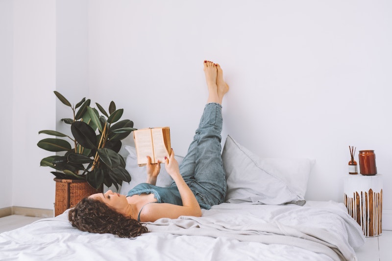 A woman reads a book in bed while stretching her legs up a wall. These believable excuses to cancel ...
