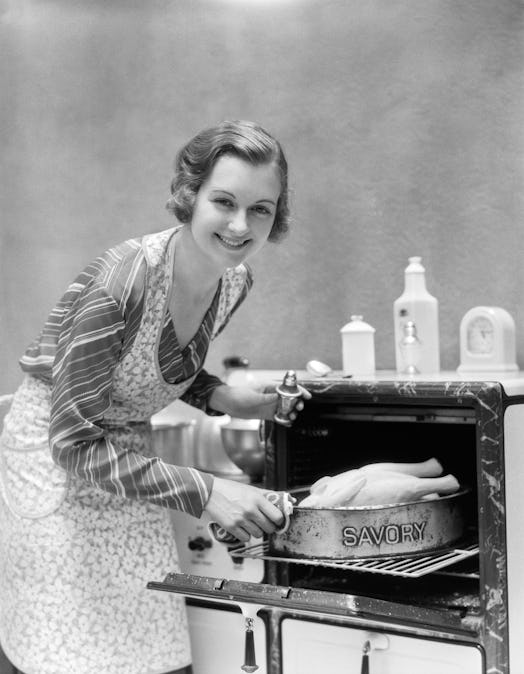 1930s HOUSEWIFE IN APRON TAKING TURKEY IN PAN READING SAVORY OUT OF OVEN LOOKING AT CAMERA  (Photo b...