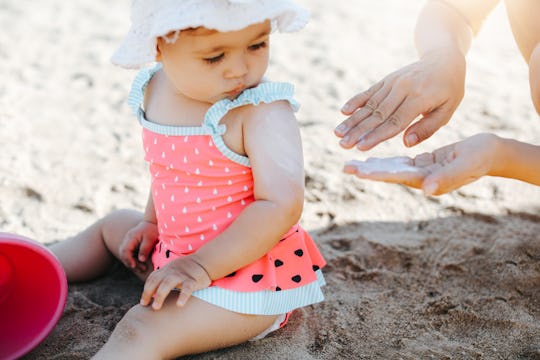 Happy laughing toddler girl having fun on sand, mom applying sunscreen