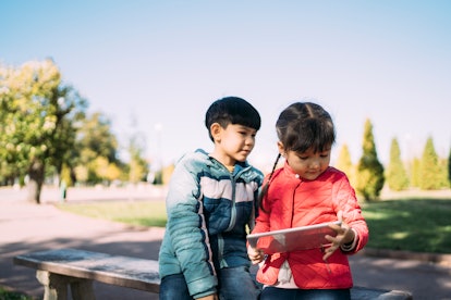 Two children looking at a iPad sitting on a bench in the park on a bright sunny day