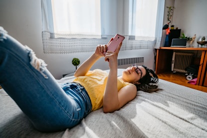 Smiling young Caucasian woman lying in bed and checking social media or reading news on smartphone.