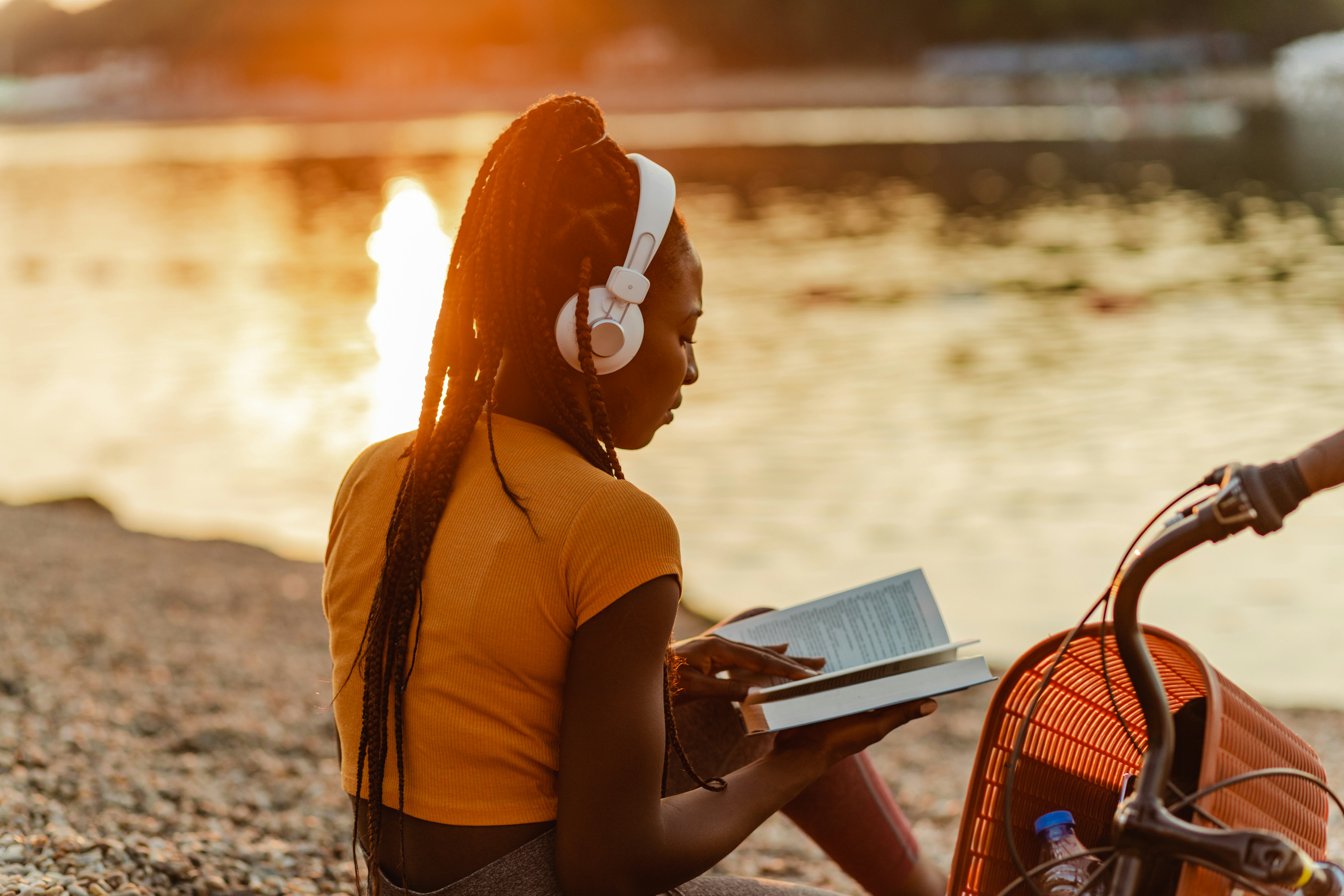 Photo of a young African American woman is using wireless headphones, she is sitting on the lakeshor...