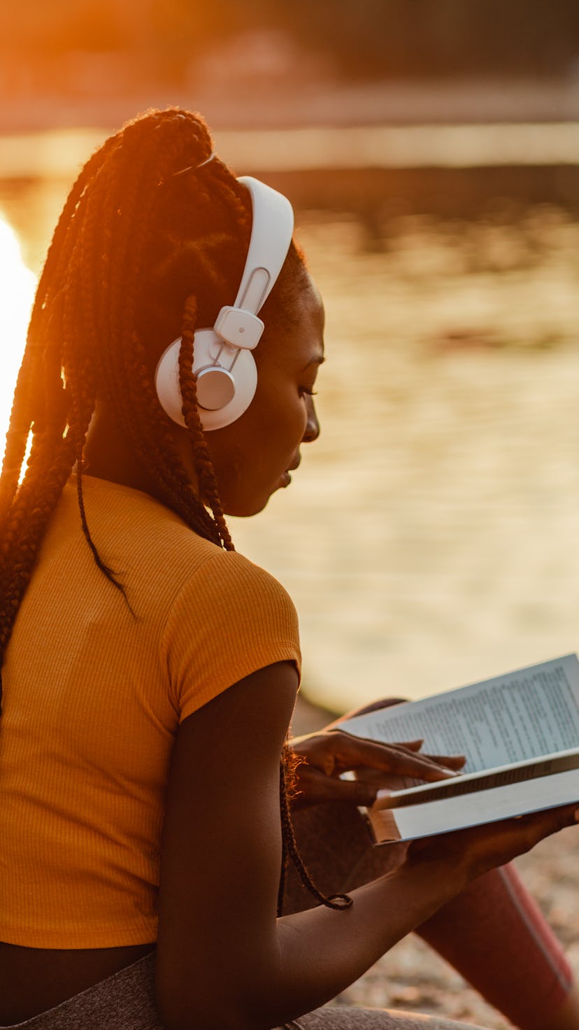 Photo of a young African American woman is using wireless headphones, she is sitting on the lakeshor...