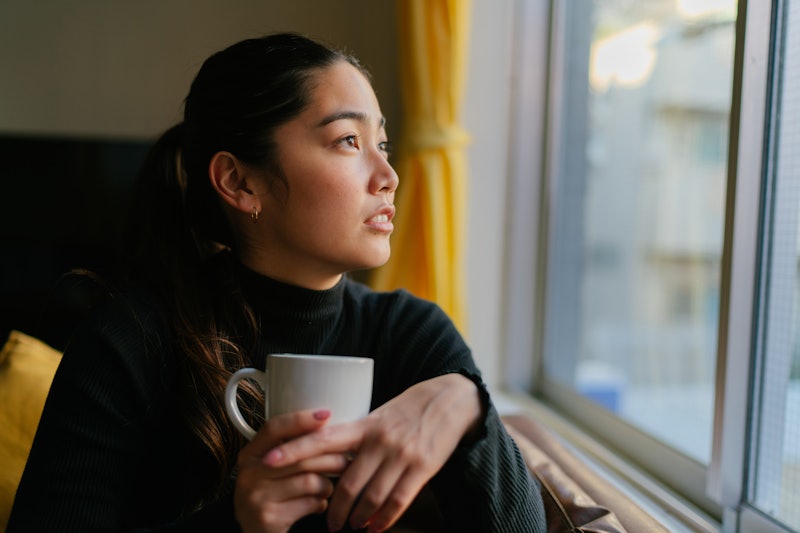A woman in a turtleneck looks out the window, stressed out after drinking a cup of coffee.