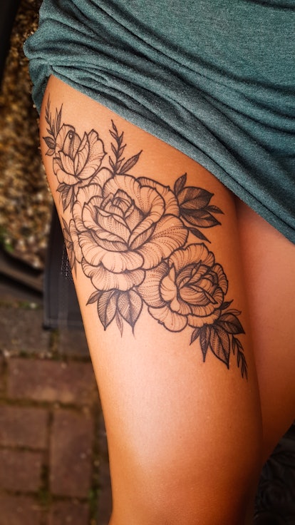 Black roses tattoo on a women's thigh part of the leg. with some leaves decoration. The woman wearin...