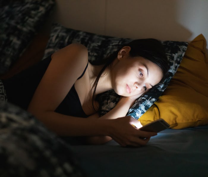 Young woman using smart phone in bed at night, staring at illuminated screen unable to fall asleep, ...