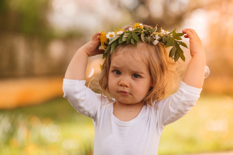 Cute baby girl standing in the park on a sunny spring day and holding her flower crown on her head w...