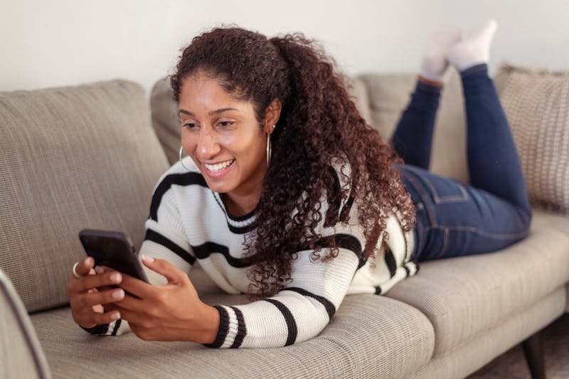 A beautiful black woman lies comfortably on her stomach on a grey couch. She is using her mobile pho...