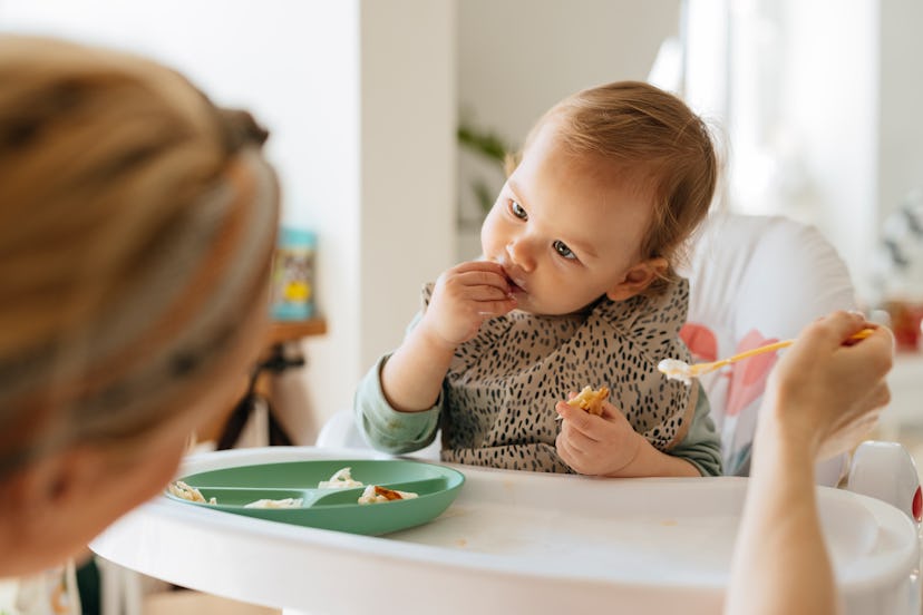 Cute baby girl with head cocked eating meal at high chair. Mother is feeding adorable toddler with s...