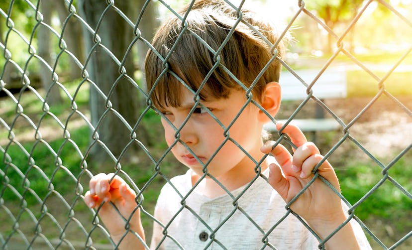 Caucasian Serious pensive Little boy looking through a schoolyard chainlink fence