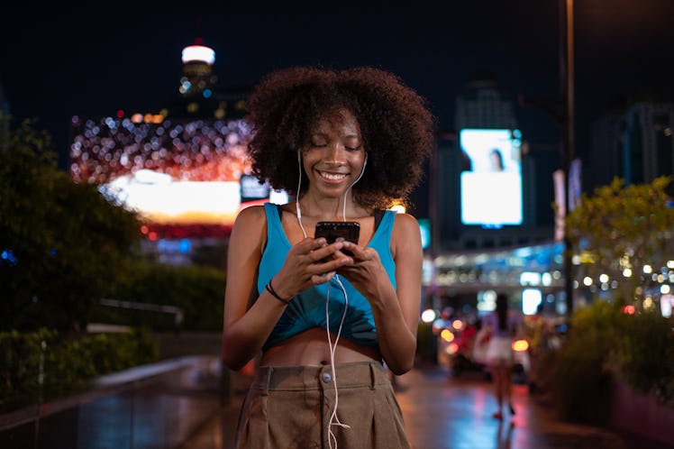 Smiling teenage girl is listening music and typing message on smart phone in the city at night.