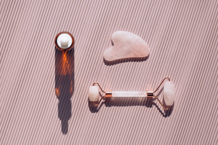 Rose quartz roller and guasha near glass bottle on striped pastel light pink background. Massagers a...