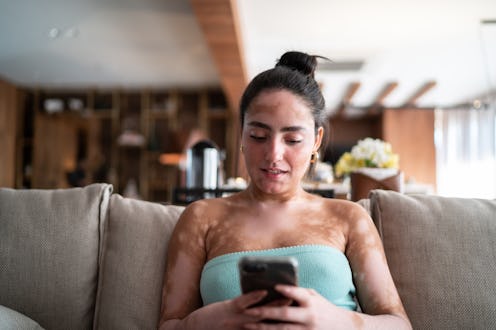 Young woman using mobile phone sitting on the couch at home to send money to Twitter friends through...