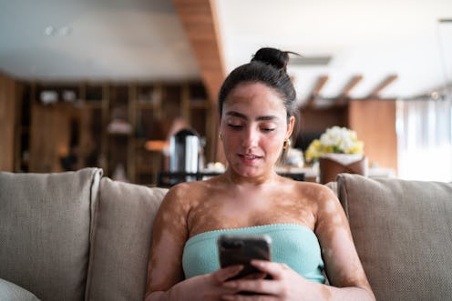 Young woman using mobile phone sitting on the couch at home to send money to Twitter friends through...