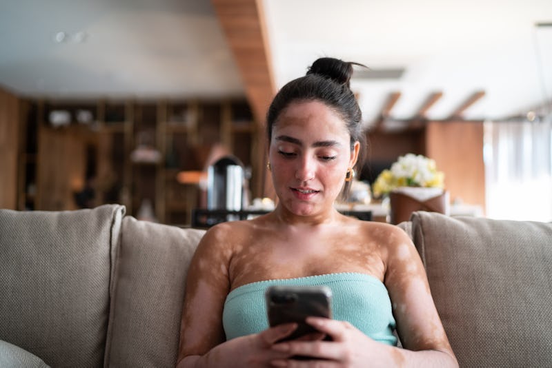 Young woman using mobile phone sitting on the couch at home to send money to Twitter friends through...