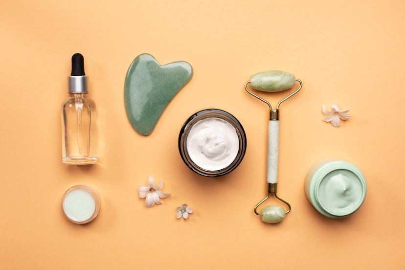 Flat lay of small flowers placed near jade spa tools and various natural cosmetic products on orange...