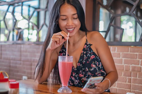 Front view shot of a smiling Indonesian woman using phone in a restaurant while waiting for her lunc...