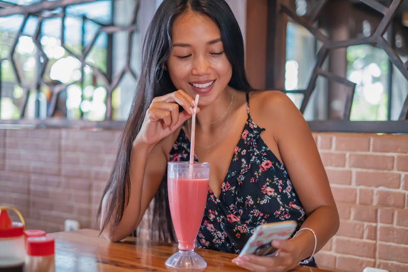 Front view shot of a smiling Indonesian woman using phone in a restaurant while waiting for her lunc...