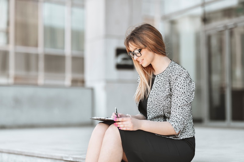 Cute Female With Eyeglasses Taking Notes On Clipboard While Relaxing In Front Of Office Building
