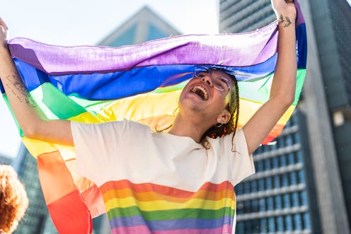 A queer person dances with multiple rainbow pride flags at a parade. Here's what the colors of diffe...