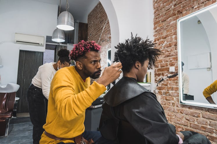 Black brazilian hairstylist with bright pink tips in his hair and wearing a yellow sweater cutting a...