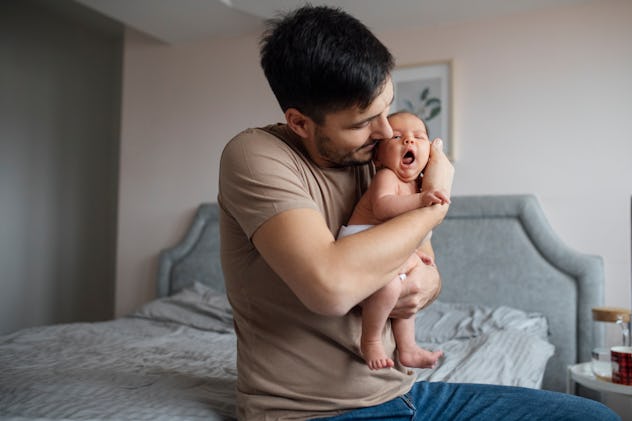 Father sitting on bed and holding his sleeping baby girl in hands kissing her
