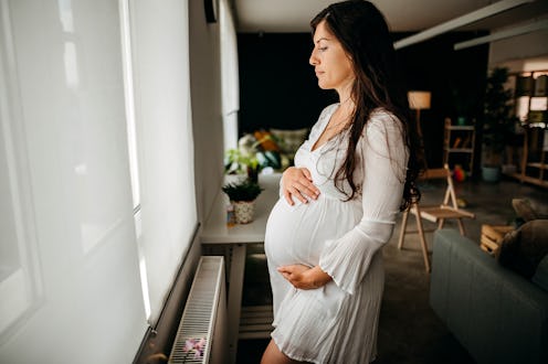 Pregnant woman standing near window and drinking hot drink