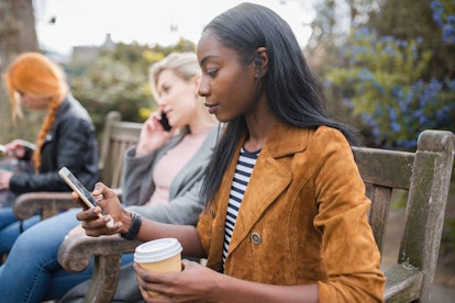 Attractive adult black woman with  friends using her phone while holding coffee cup in public park i...