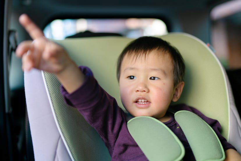 A small baby boy is sitting in a safety seat of a rear seat on a car, pointing out the window.