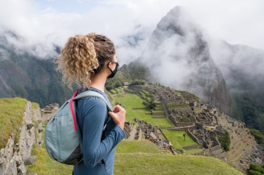 A woman wearing a face mask looks at Machu Picchu from atop a hill.