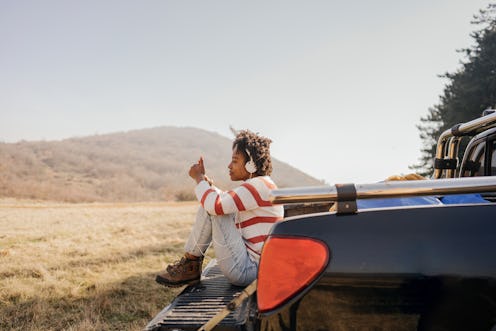 Photo of a young woman on a road trip sitting on pick-up truck texting her family to stay in touch.