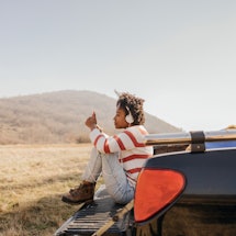 Photo of a young woman on a road trip sitting on pick-up truck texting her family to stay in touch.