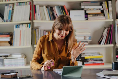 A smiling blonde businesswoman sitting at the office, eating a protein bar while video calling with ...