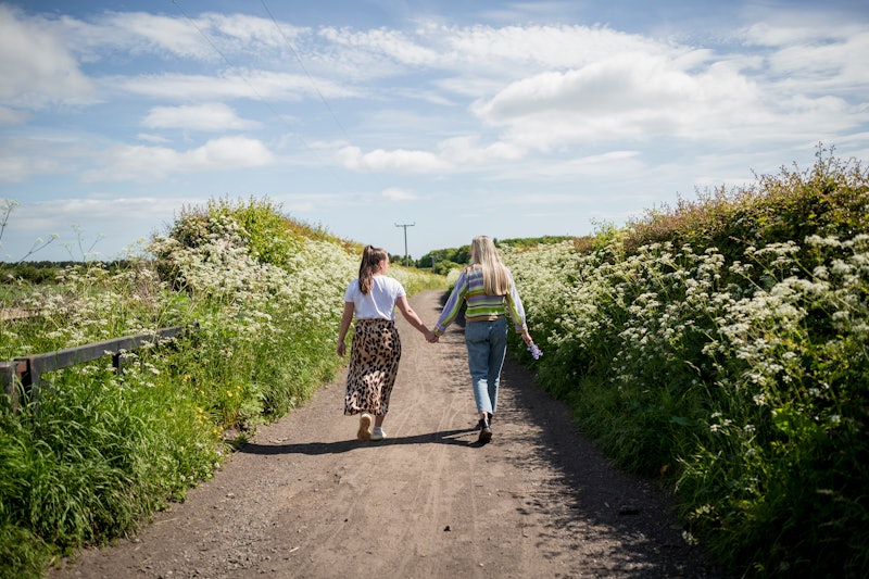 A rear-view shot of an LGBTQIA female couple walking down a country footpath together, they are wear...