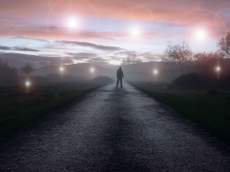 A UFO concept of glowing orbs, floating above a misty winters road just after sunset. With a silhoue...