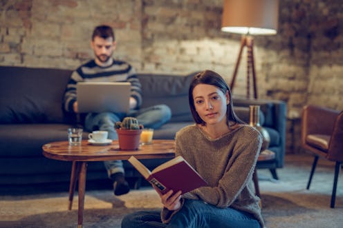 Young woman sitting on floor reading a book while young man is using laptop in the background. Looki...