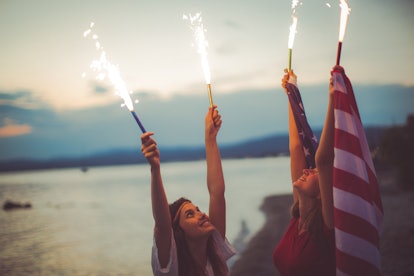 Two girls holding sprinkles and waving an American flag at the beach as a way to celebrate fourth of...