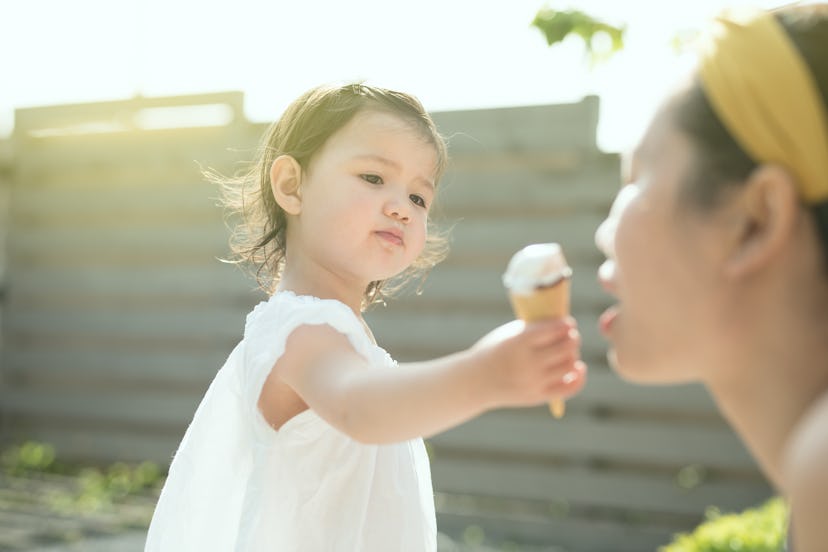 A toddler wearing a white dress offers an ice cream cone to her mother in a sunny spring day in the ...