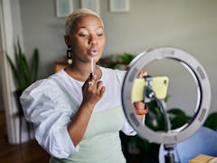 Young African female influencer showing how to apply lip gloss while doing a vlog post from home usi...