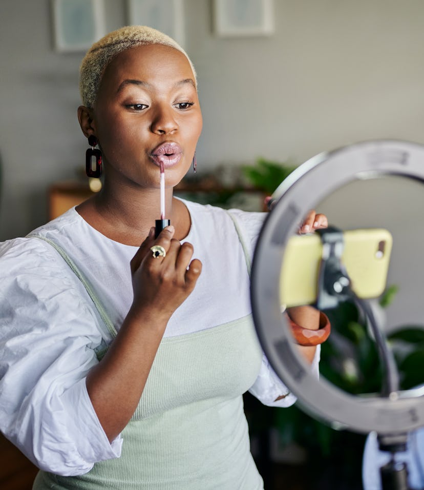 Young African female influencer showing how to apply lip gloss while doing a vlog post from home usi...