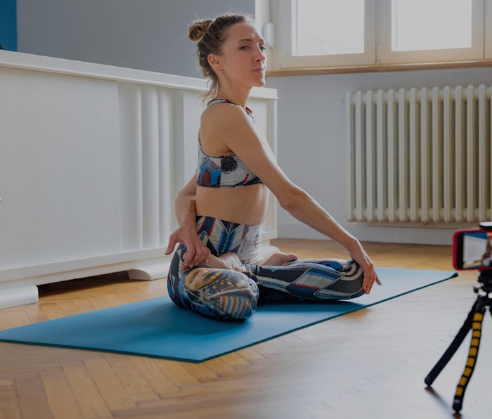 Woman recording yoga class tutorial, sitting in lotus pose on exercise mat at home with left arm beh...