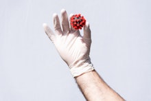 Man's hand wearing a disposable white protective glove, holding a coronavirus model with first finge...