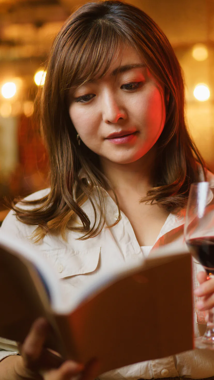 A woman is enjoying reading a book and drinking red wine in a cozy comfortable cafe & bar at night.