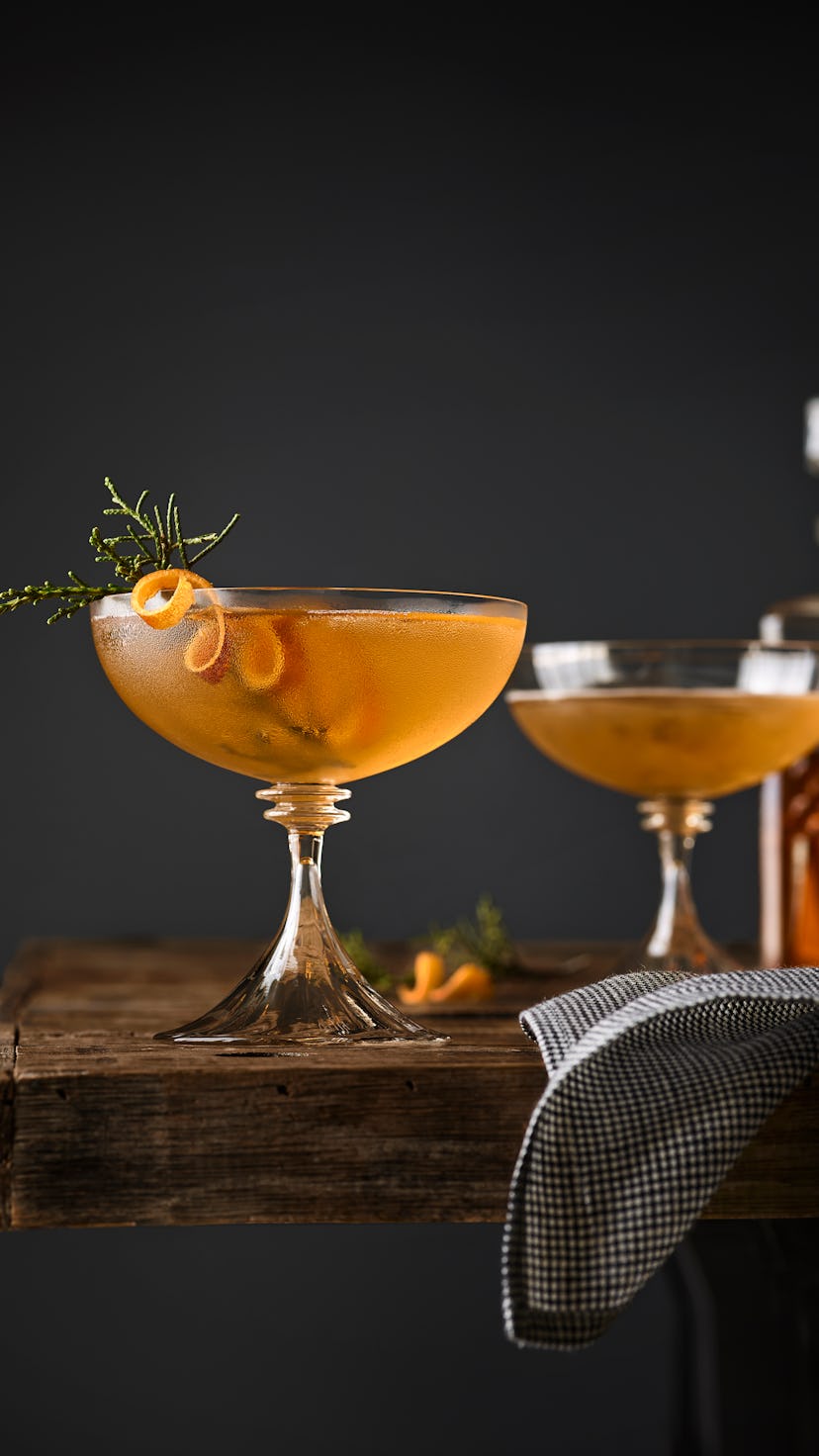 Still life of two Manhattan cocktails garnished with Juniper on a table with whiskey decanter in the...